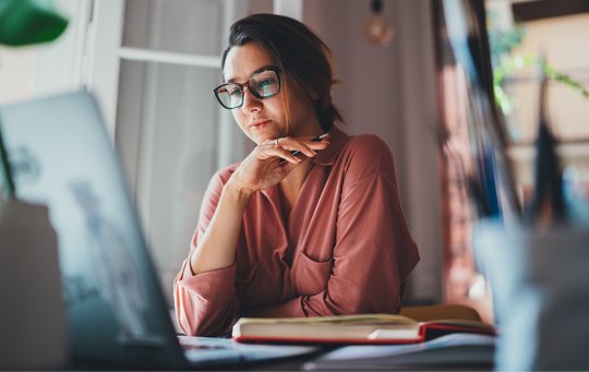 Woman wearing glasses, holding a pen sitting at a table with open books and a laptop